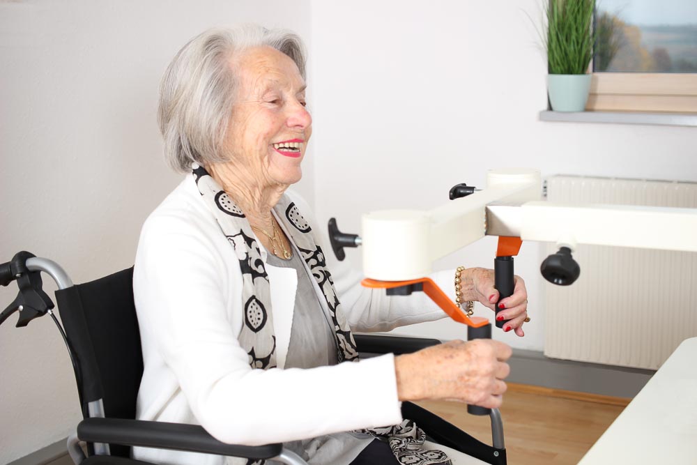 Seniorin mit grauen Haaren in einem Rollstuhl sitzt lachend an der Drehorgel des Plaudertischs.